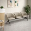 Longarina de 3 lugares modelo Nantes com acabamento em couro sintético fendi, ambientada em uma sala de espera moderna com balcão de atendimento, plantas e quadros decorativos.